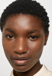 Gros plan sur un visage avec des cheveux noirs courts et bouclés, une peau marron lisse, un maquillage naturel, et un col de couleur neutre visible au niveau du cou.