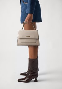 Beige struktureret håndtaske med en top håndtag, glat læder og guldfarvet hardware, kombineret med en tætsiddende blå jakke og brune knælange støvler.