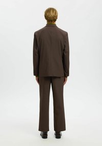 Pruun kohandatud ülikond, millel on istuv jakk ja sirge säärega püksid. Sile kangapind, minimalistlikud detailid ja ühtlane tone kogu ulatuses.