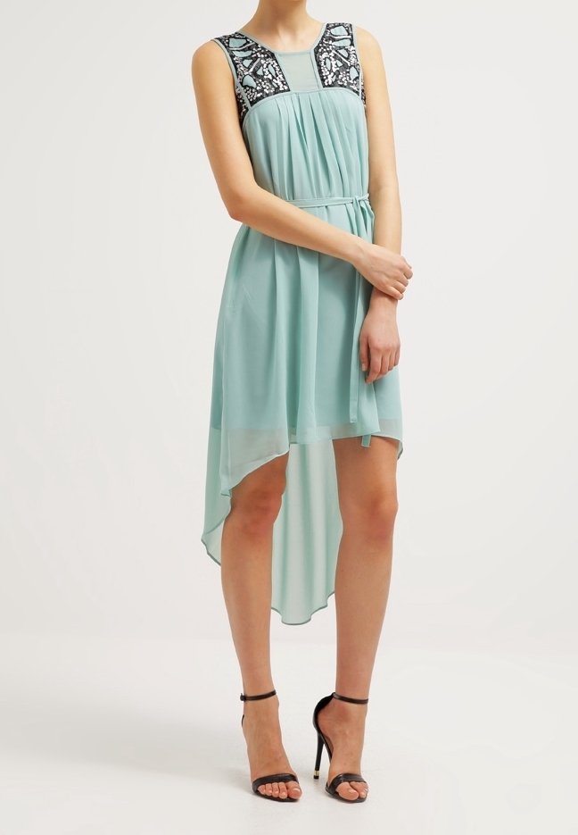 Femme portant une robe bleu clair sans manches à jupe asymétrique avec un yoke à motif noir et blanc, associée à des escarpins noirs à bride à la cheville.