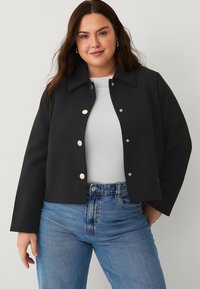 Black cropped jacket with large collar, featuring gold snap buttons. Worn over a white shirt with blue denim jeans. Smooth texture, structured fit.
