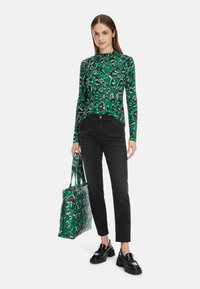 Grünes Langarmoberteil mit Leopardenmuster, schwarze Hose und klobige schwarze Slipper. Tragend eine passende grüne Tasche mit ähnlichem Druckdesign.