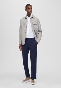 Giacca button-up grigio chiaro con due taschini sul petto, camicia bianca sotto, pantaloni navy e sneakers beidge. Design e tessuto semplici.
