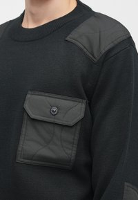 Melnā puloveri džemperis ar teksturētu materiālu, kuram ir quilted plecu akcenti un krūšu kabata ar pogas aizdari.