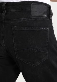 Jeans de mezclilla negra con un acabado texturizado, costuras en contraste y un bolsillo trasero con un pequeño detalle de logo bordado.