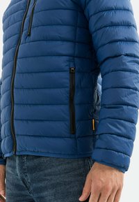 Blå pufferjakke lavet af glat, skinnende materiale. Har vandret quiltning, en sort lynlås og en sidelomme med en kontrastfyldt gul accent.