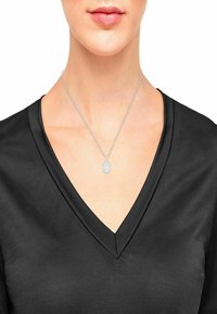 Collana con pendente in argento dal design circolare, con un centro texture, indossata su una catena fine, contro una maglietta nera a V.
