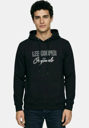 Jeune homme aux cheveux bouclés portant un sweat à capuche noir avec le texte « LEE COOPER Originals » et un jean bleu foncé, debout devant un fond uni.