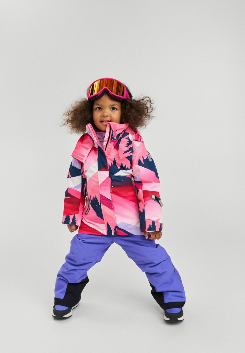 Farverig skijakke med et pink og blåt abstrakt mønster, båret over et par lilla skibukser. Inkluderer hætte og lynlåselementer.