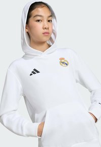 Sweat à capuche blanc avec une poche frontale, trois bandes noires Adidas et un petit logo du Real Madrid sur la poitrine ; le tissu semble lisse et épais.