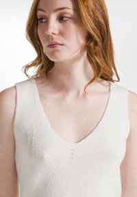 Top de tank sin mangas de punto acanalado blanco con un profundo escote en V. La tela parece suave y texturizada, con detalles sutiles de encaje en el escote.