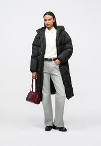 Mulher vestindo um longo casaco acolchoado preto, suéter branco, jeans cinza claro, sapatos pretos e segurando uma bolsa borgonha.