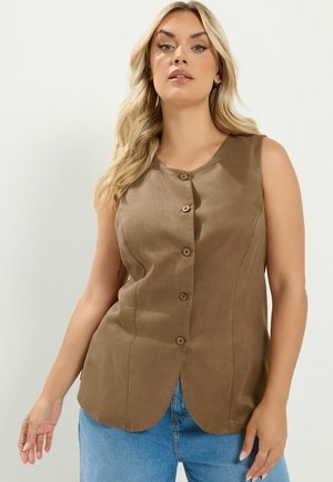 Kvinde med langt blond hår iført brunærmeløs knapbluse og blå jeans mod en ensfarvet lys baggrund.