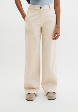 Person, die beige weite Hosen mit Taschen, ein hellblaues Oberteil und beige Slip-On-Schuhe trägt, steht vor weißem Hintergrund.