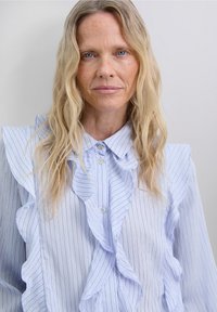 Blonde woman with long wavy hair wearing a light blue pinstriped blouse with ruffled collar and front detailing, neutral background.