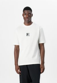 Hvid T-shirt med korte ærmer lavet af bomuld, med et lille gråt Jordan-logo i midten, afslappet pasform og rund hals. 