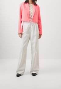 Giacca rosa cropped sopra un top in pizzo bianco, abbinata a pantaloni bianchi a vita alta e scarpe nere. Tessuto morbido con una vestibilità rilassata.