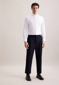 Wit overhemd met lange mouwen en knoopsluiting, gecombineerd met marineblauwe geplooide broek. Zwarte nette schoenen maken de outfit compleet, tegen een neutrale achtergrond.