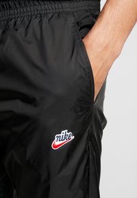 Mustat nailoniset urheiluhousut, joissa joustava vyötärönauha, sivutasku ja valkoinen, punainen sekä sininen Nike-logo kirjailtuna.