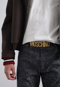 Musträbel mustriga hallide pükste ja tumeda tõmbluku jakiga, millel on kuldne "MOSCHINO" kiri.
