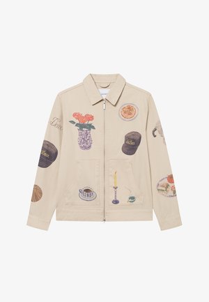 Beige canvas jacket featuring various artistic prints including flowers, food, and household items. Zipper front and two front pockets.