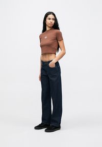 Γυναίκα που στέκεται με το ένα χέρι στην τσέπη, φοράει καφέ κοντομάνικη μπλούζα, σκούρο μπλε τζιν και μαύρα αθλητικά παπούτσια σε λευκό φόντο.