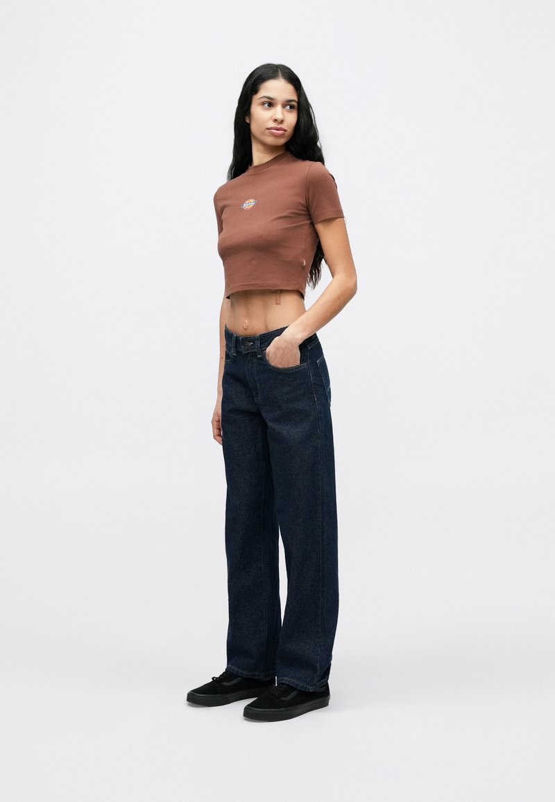 Femme debout, une main dans la poche, portant un t-shirt marron court, un jean bleu foncé et des baskets noires sur fond blanc.