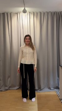 Γυναίκα στέκεται ξυπόλητη σε ξύλινο πάτωμα με εκρού μπλούζα μακρυμάνικη και μαύρο παντελόνι, με ανοιχτογκρι κουρτίνες στο φόντο.