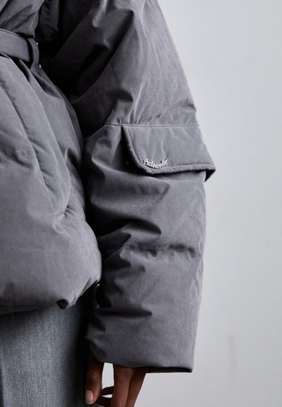 Gray puffer jacket featuring a textured fabric, wide sleeves, and a flap pocket with stitched branding. The design emphasizes warmth and coverage.