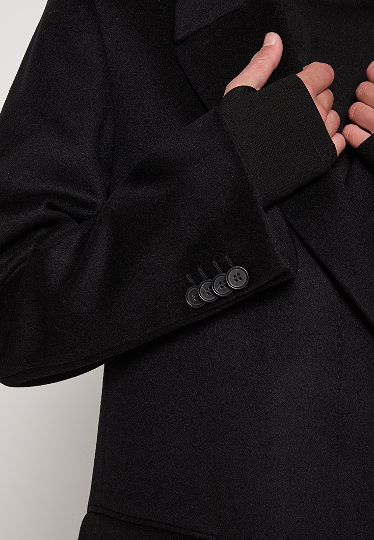 Blazer en laine noire avec un revers cranté, des manches resserrées avec des évents pour les pouces, et quatre boutons noirs aux poignets. Texture lisse.