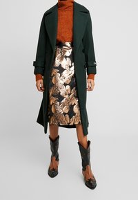 Tummanvihreä trenssitakki, ruosteenvärinen pooloneule, metallinhohtoinen kultakuvioinen hame, mustat ja ruskeat cowboy-saappaat tähtikoristein.