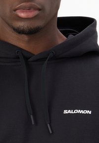 Sweatshirt à capuche noire en tissu doux, doté d'une poche kangourou à l'avant et d'un logo "SALOMON" blanc sur la poitrine.