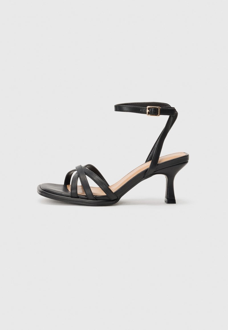 Sandale à talons noire avec lanières et bride de cheville, boucle dorée, talon courbé de hauteur moyenne, sur un fond gris clair uni.