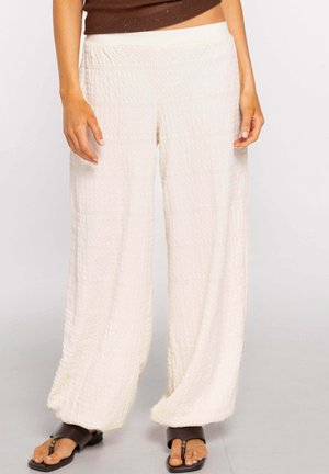 Mujer con pantalones blancos anchos y texturizados y sandalias marrones tipo 'thong' de goma, de pie sobre un fondo liso.
