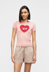 Γυναίκα που φοράει στενό ροζ t-shirt με κόκκινη καρδιά και λευκό σχέδιο λογότυπου, συνδυασμένο με μπεζ καθημερινές βερμούδες, στέκεται μπροστά σε απλό φόντο.