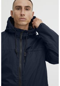 Navy blue hooded jacket with a zip front, adjustable drawstrings, and two front pockets. Made of lightweight, textured fabric.