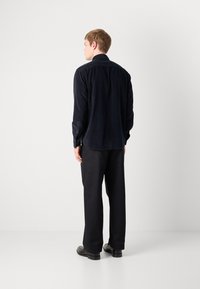 Hombre de pie de espaldas, vistiendo una camisa negra de manga larga, pantalones anchos negros y zapatos de vestir negros, frente a un fondo blanco.