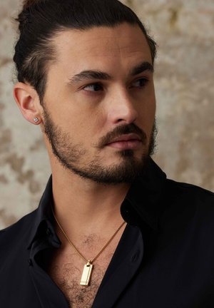 Collier en pendentif doré sur une chaîne fine, porté sur une peau nue, avec une chemise noire et une boucle d'oreille visible, mettant en valeur les textures et les formes.