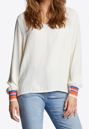 Femme portant un chemisier blanc à manches longues avec des poignets rayés multicolores et un jean bleu clair avec de petits motifs floraux.