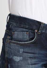 Lähedane tumblue denim teksapüksid, millel on tuhmunud kohad ja väike esikott, kantud valge särgi sisse tõmmatud.