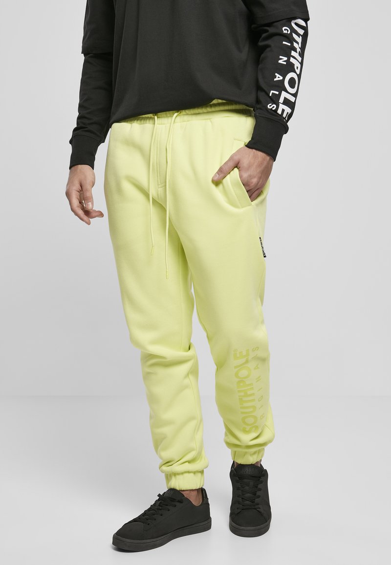 Pantaloni de trening galbeni neon cu manșete elastice, talie cu șnur și buzunare laterale. Prezintă logo-ul "SOUTHPOLE" imprimat vertical. O pereche de adidași negri.