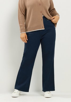 Persoon die een bruine, geknoopte cardigan, donkerblauwe wijde broek en witte sneakers draagt, staande tegen een eenvoudige lichte muur.