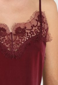 Camisola bordeaux con scollo in pizzo, caratterizzata da motivi floreali, bordi frangiati e spalline sottili. Tessuto morbido con una superficie liscia.