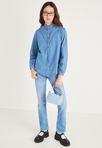 Denimskjorte med knapper og volangkant, kombinert med lyseblå jeans og en liten lyseblå håndveske. Kraftige svarte platåsko.