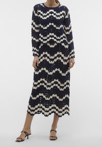 Abito in crochet blu navy a maniche lunghe, con motivi ondulati color crema, vestibilità comoda e lunghezza media. Indossato con sandali neri con fascia.