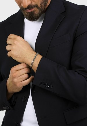 Blazer nero con vestibilità slim e rever a punta, indossato su una camicia bianca. Presenta un braccialetto d'argento al polso che tiene il blazer.