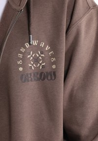 Felpa con zip marrone realizzata in tessuto morbido. Presenta il logo "SANDWAVES" sopra un motivo floreale e "Oxbow" in lettere in grassetto nella parte inferiore.