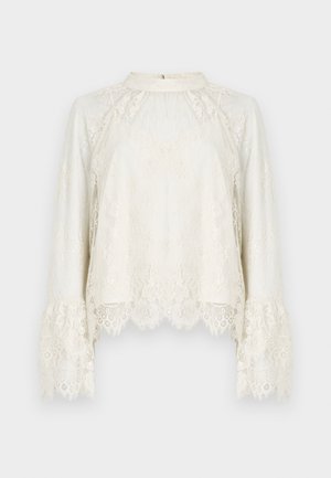 Top din dantelă în nuanță crem, cu mâneci lungi și evazate, și un tiv cu margini ondulate. Prezintă modele florale intricate și un decolteu înalt, rotund.