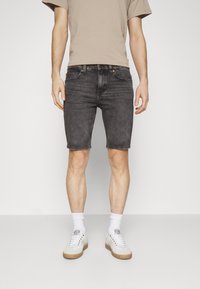 Modellen står framför en enkel bakgrund, klädd i en beige t-shirt, mörk denimshorts och vita sneakers med synliga ankelsockar.