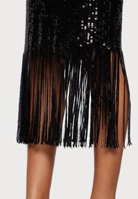 Černé šaty se flitry a třásněmi na lemu. Materiál je lesklý s perlovým vzhledem, s bočními rozparky pro větší pohyb.